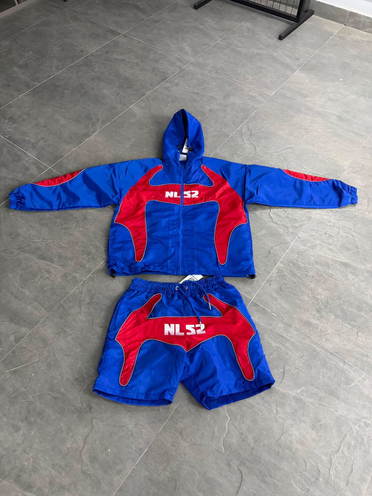 A two-piece set consisting of a hooded zip-up jacket and matching shorts, laid flat on a gray tiled floor. The set features a vibrant blue base with red graphic panels and white 'NLS2' lettering on the chest and shorts.