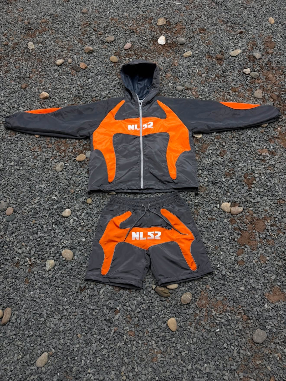 A dark gray and bright orange windbreaker jacket and matching shorts laid flat on a gravel surface. The jacket features a hood, a front zipper, and the text 'NLS2' printed in white on the chest, while the shorts also display 'NLS2' on the front.
